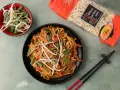 Pyszny makaron z mięsem i warzywami, posypany sezamem. Obok miseczka kiełków fasoli mung i paczka makaronu chow mein.
