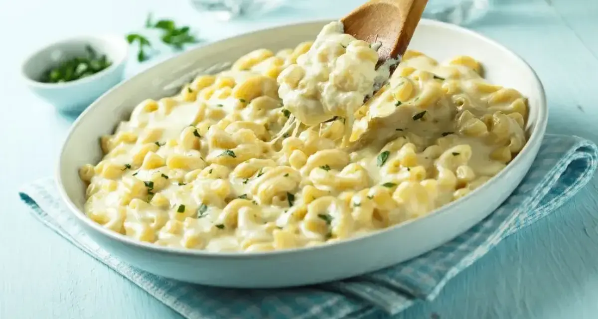 Pyszny makaron z mozzarellą i ziołami, podany w białej misce, z drewnianej łyżki nakładana jest porcja ciągnącego się sera.