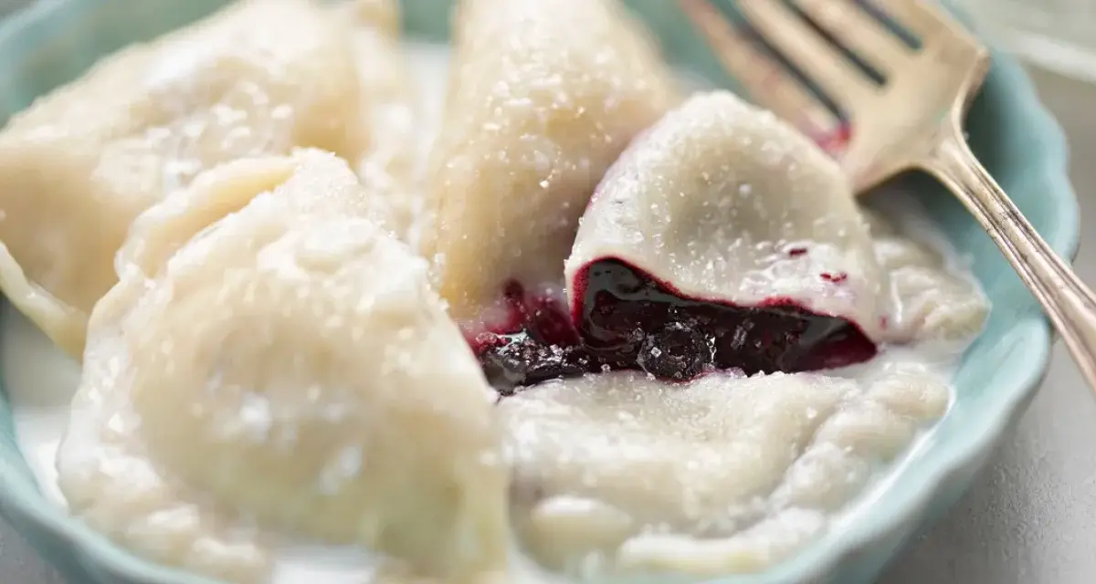 Pyszne pierogi z jagodami, polane śmietaną, w niebieskiej miseczce. Widoczny widelec.