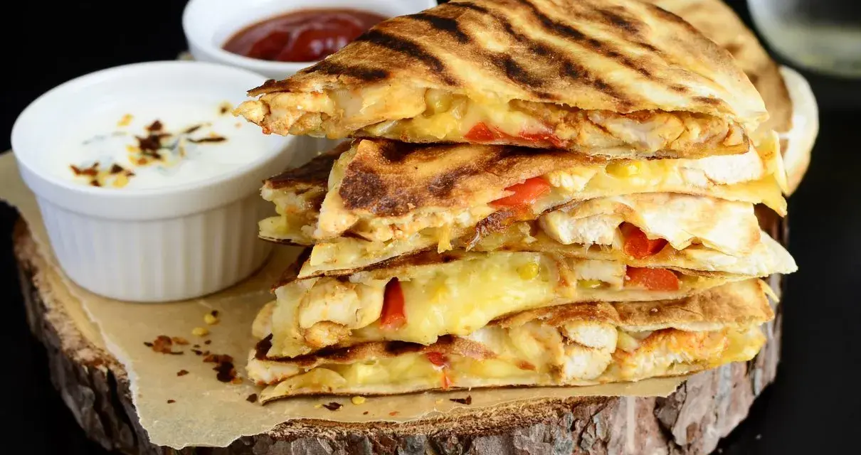 Stos pysznych kawałków quesadilli z kurczakiem, roztopionym serem i papryką, podane z dipami.