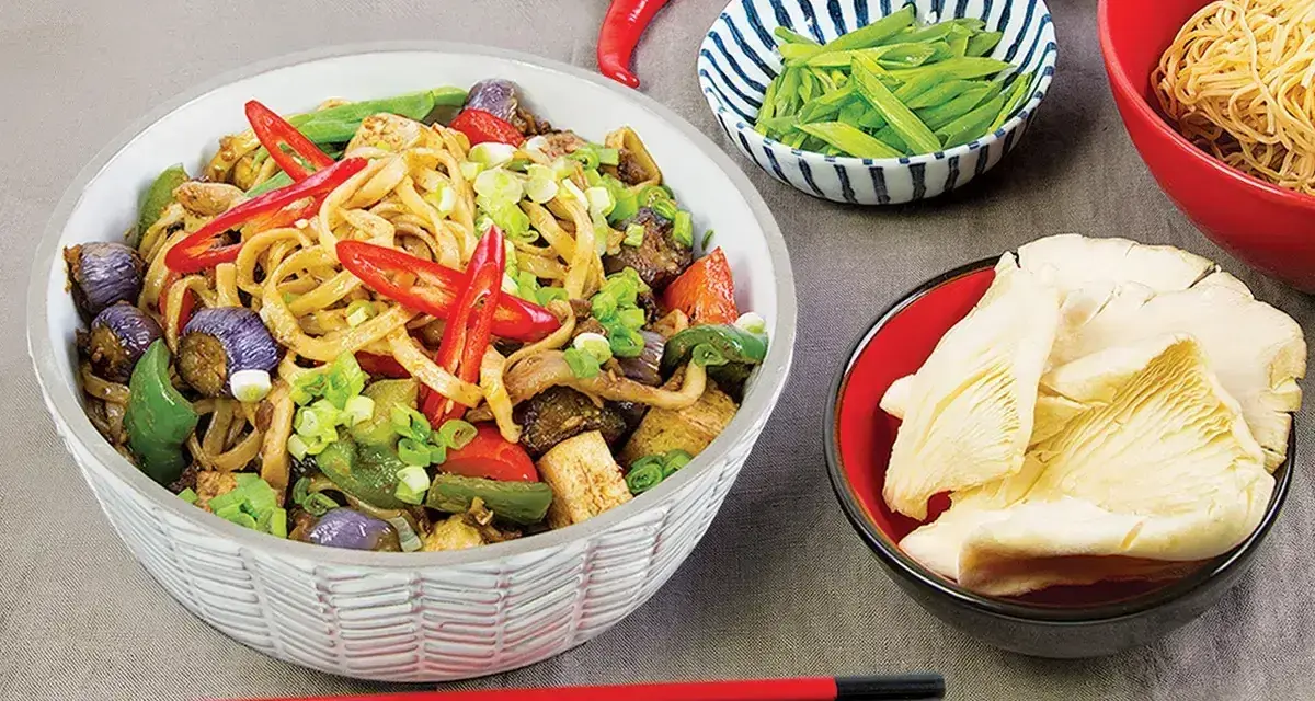 Pyszny makaron chow mein z warzywami i papryczkami chili, obok miseczki z grzybami i fasolką.
