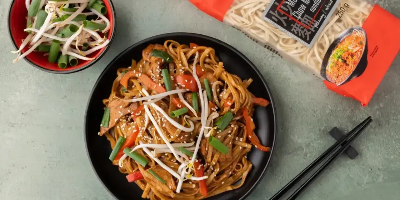 Pyszny makaron z mięsem i warzywami, posypany sezamem. Obok miseczka kiełków fasoli mung i paczka makaronu chow mein.