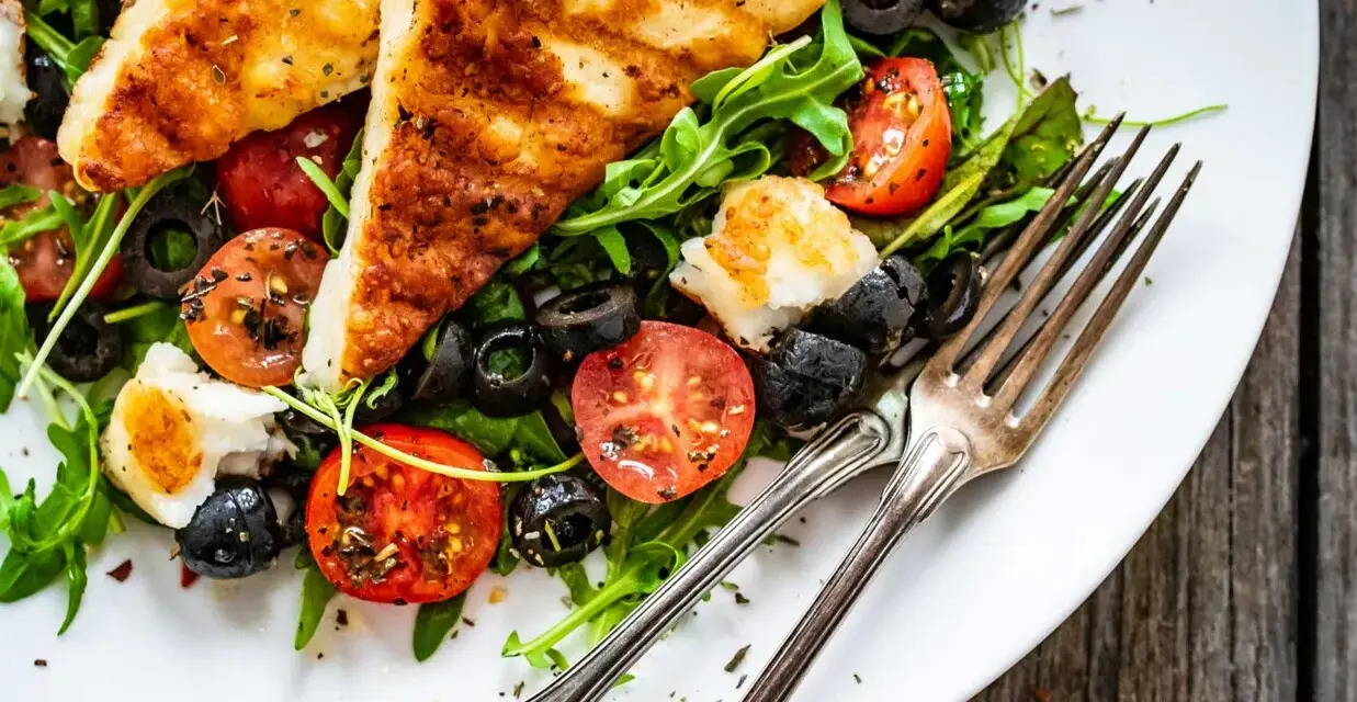 Pyszna sałatka z grillowanym halloumi, pomidorkami koktajlowymi i czarnymi oliwkami na rukoli. Idealna na lekki posiłek.