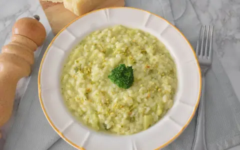 Kremowe risotto z brokułami, posypane pistacjami, z małą różyczką brokułu na wierzchu. Obok widelca i kawałków parmezanu.