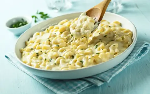 Pyszny makaron z mozzarellą i ziołami, podany w białej misce, z drewnianej łyżki nakładana jest porcja ciągnącego się sera.