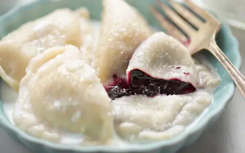 Pyszne pierogi z jagodami, polane śmietaną, w niebieskiej miseczce. Widoczny widelec.