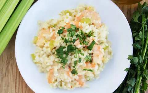 Kremowe risotto z krewetkami, ozdobione natką pietruszki, obok łodyg selera i oliwy.