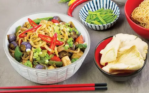 Pyszny makaron chow mein z warzywami i papryczkami chili, obok miseczki z grzybami i fasolką.