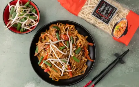 Pyszny makaron z mięsem i warzywami, posypany sezamem. Obok miseczka kiełków fasoli mung i paczka makaronu chow mein.