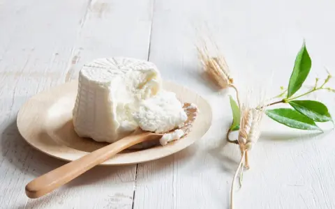 Świeża ricotta na drewnianej łyżce, obok kłosy zbóż i zielone gałązki. Idealna do dań, gdy szukasz czym zastąpić ricottę.