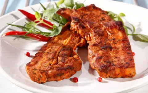 Dwa soczyste kawałki boczku na grilla, przyprawione i ozdobione papryczkami chili i zielonymi listkami.