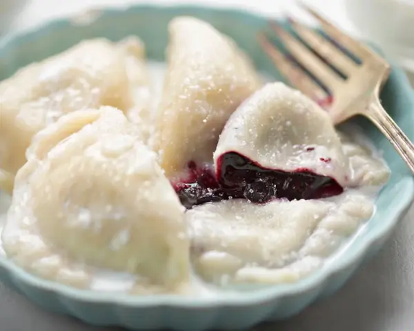 Pyszne pierogi z jagodami, polane śmietaną, w niebieskiej miseczce. Widoczny widelec.