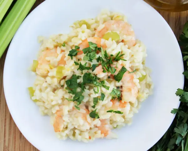 Kremowe risotto z krewetkami, ozdobione natką pietruszki, obok łodyg selera i oliwy.