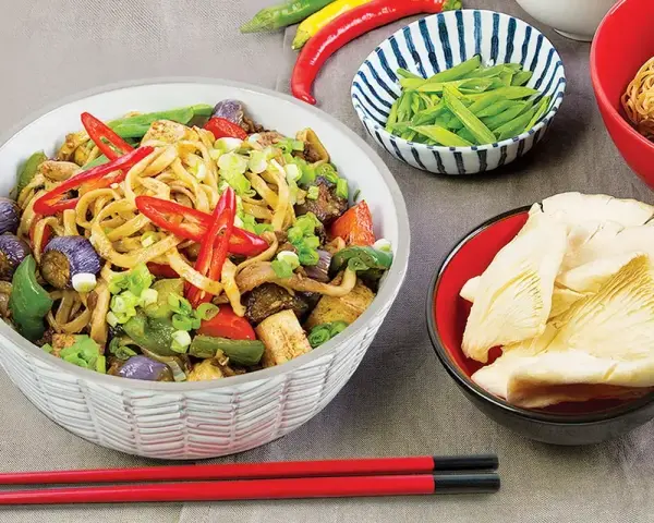 Pyszny makaron chow mein z warzywami i papryczkami chili, obok miseczki z grzybami i fasolką.