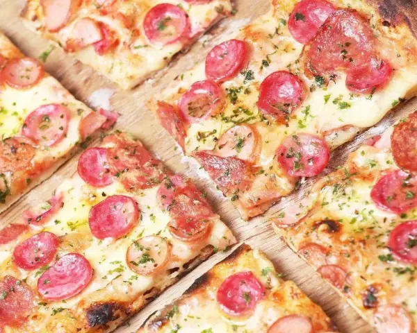 Kawałki pysznej pizzy na cienkim cieście z kiełbasą i serem, posypane ziołami.