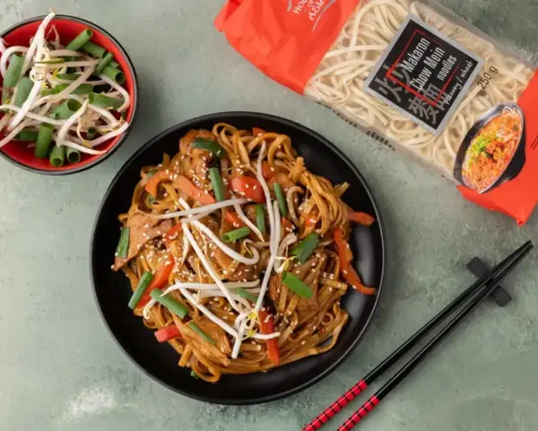 Pyszny makaron z mięsem i warzywami, posypany sezamem. Obok miseczka kiełków fasoli mung i paczka makaronu chow mein.