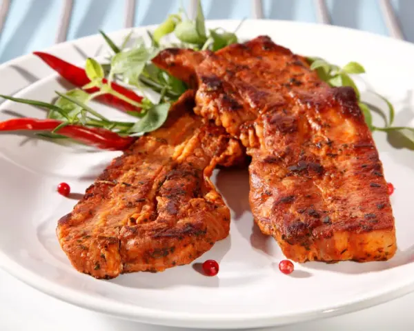 Dwa soczyste kawałki boczku na grilla, przyprawione i ozdobione papryczkami chili i zielonymi listkami.