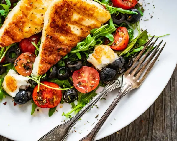 Pyszna sałatka z grillowanym halloumi, pomidorkami koktajlowymi i czarnymi oliwkami na rukoli. Idealna na lekki posiłek.