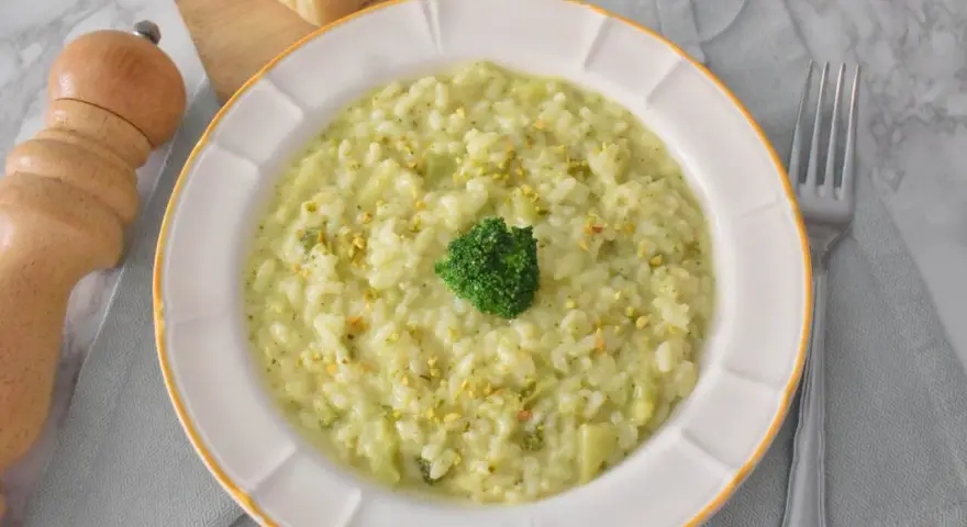 Kremowe risotto z brokułami, posypane pistacjami, z małą różyczką brokułu na wierzchu. Obok widelca i kawałków parmezanu.