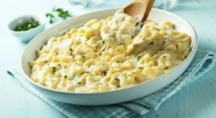 Pyszny makaron z mozzarellą i ziołami, podany w białej misce, z drewnianej łyżki nakładana jest porcja ciągnącego się sera.