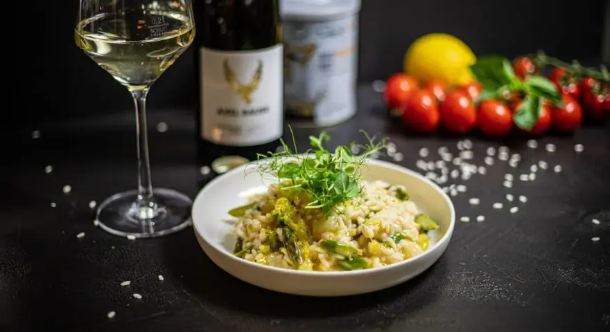 Aksamitne risotto z kurczakiem i szparagami, udekorowane kiełkami. Obok kieliszek białego wina i butelka.