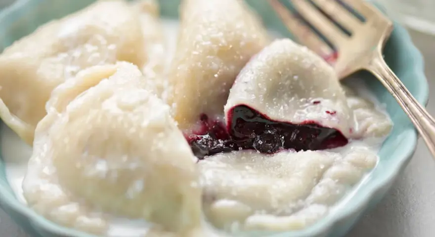 Pyszne pierogi z jagodami, polane śmietaną, w niebieskiej miseczce. Widoczny widelec.