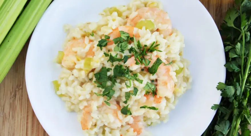 Kremowe risotto z krewetkami, ozdobione natką pietruszki, obok łodyg selera i oliwy.