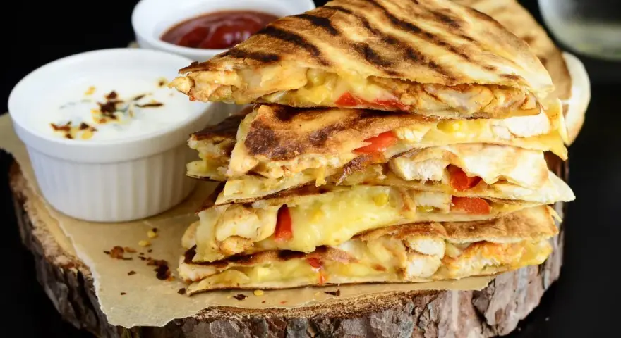 Stos pysznych kawałków quesadilli z kurczakiem, roztopionym serem i papryką, podane z dipami.
