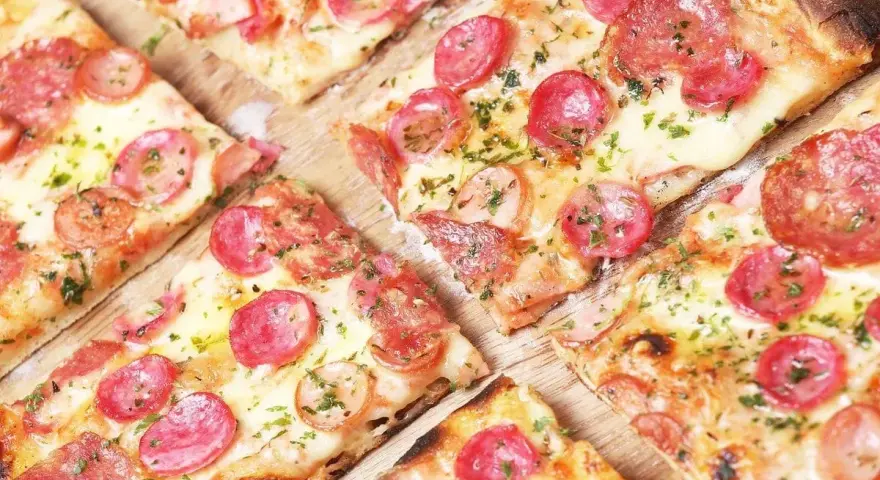 Kawałki pysznej pizzy na cienkim cieście z kiełbasą i serem, posypane ziołami.