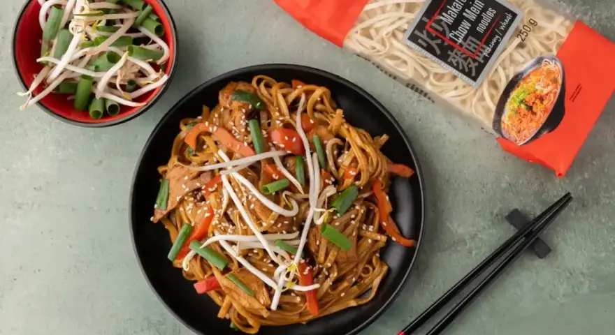 Pyszny makaron z mięsem i warzywami, posypany sezamem. Obok miseczka kiełków fasoli mung i paczka makaronu chow mein.