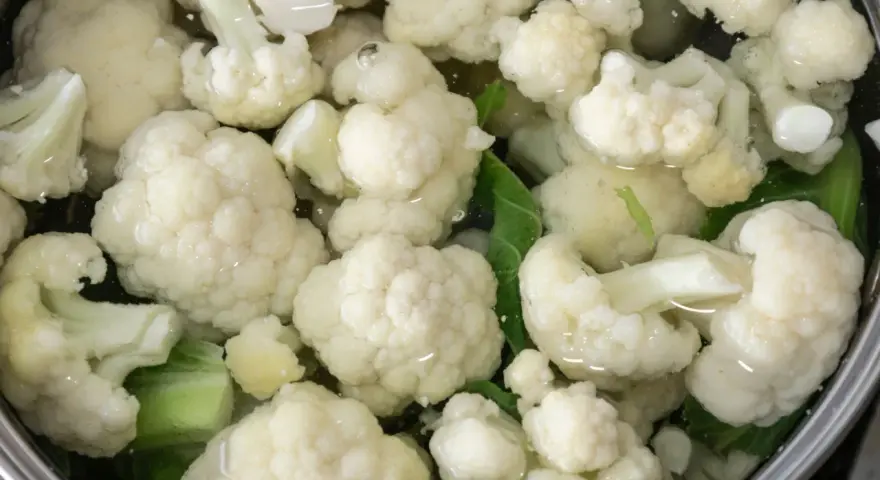 Kalafior gotuje się w garnku z wodą. Białe różyczki i zielone liście są zanurzone w płynie, tworząc apetyczny widok.