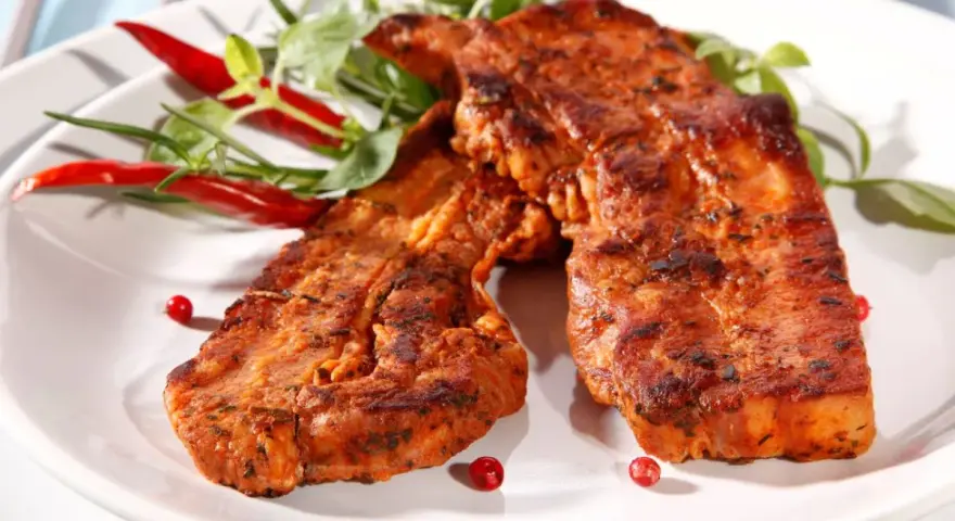 Dwa soczyste kawałki boczku na grilla, przyprawione i ozdobione papryczkami chili i zielonymi listkami.