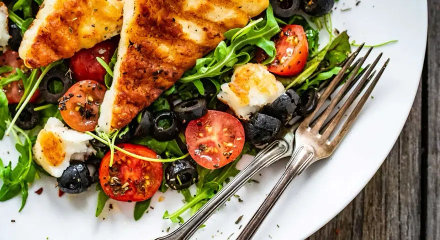 Pyszna sałatka z grillowanym halloumi, pomidorkami koktajlowymi i czarnymi oliwkami na rukoli. Idealna na lekki posiłek.