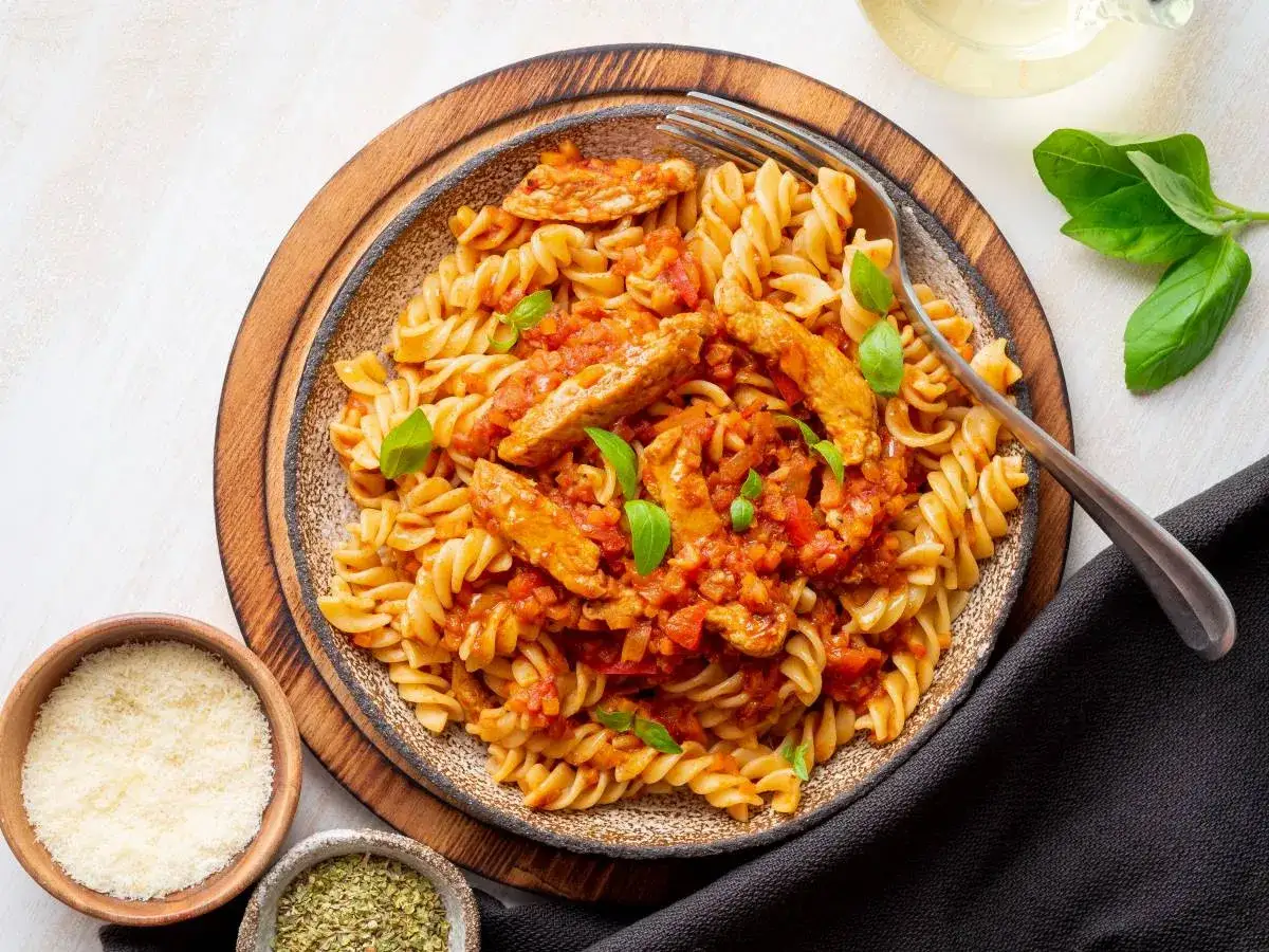Szybki obiad z makaronem fusilli w sosie pomidorowym z kawałkami kurczaka i świeżą bazylią. Obok miseczki z parmezanem i ziołami.