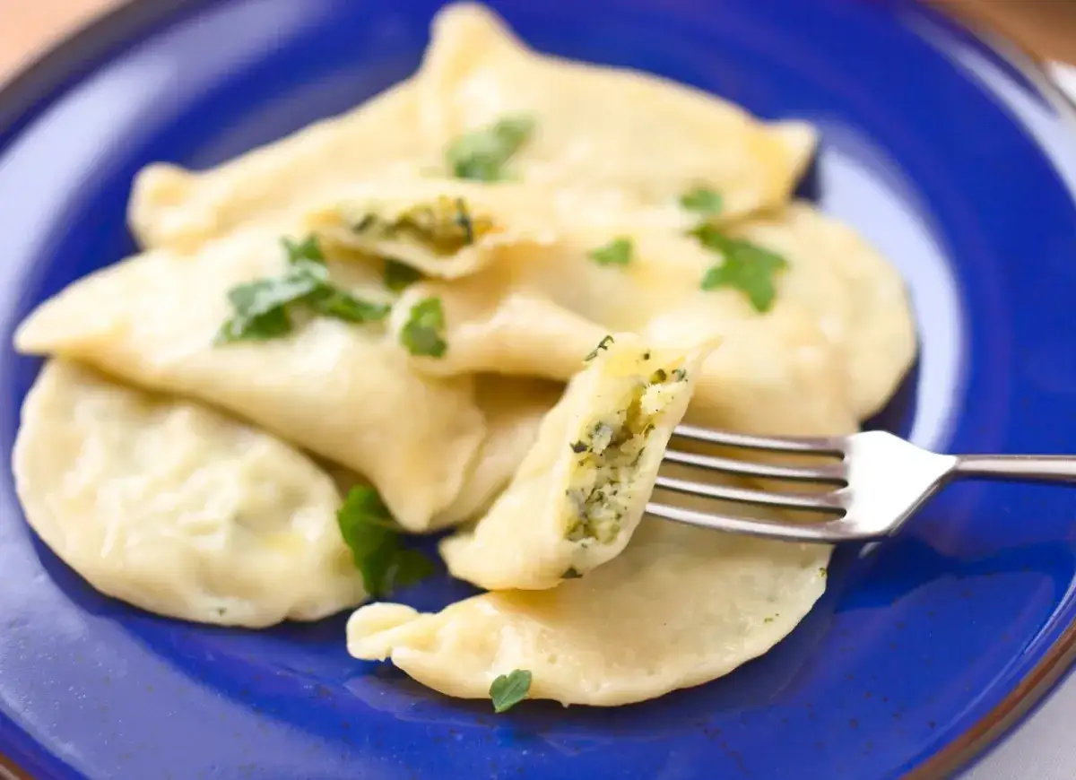 Pyszne pierogi ze szpinakiem i serem, udekorowane natką pietruszki, na niebieskim talerzu. Jedna pierożka jest podniesiona widelcem, ukazując bogate nadzienie.