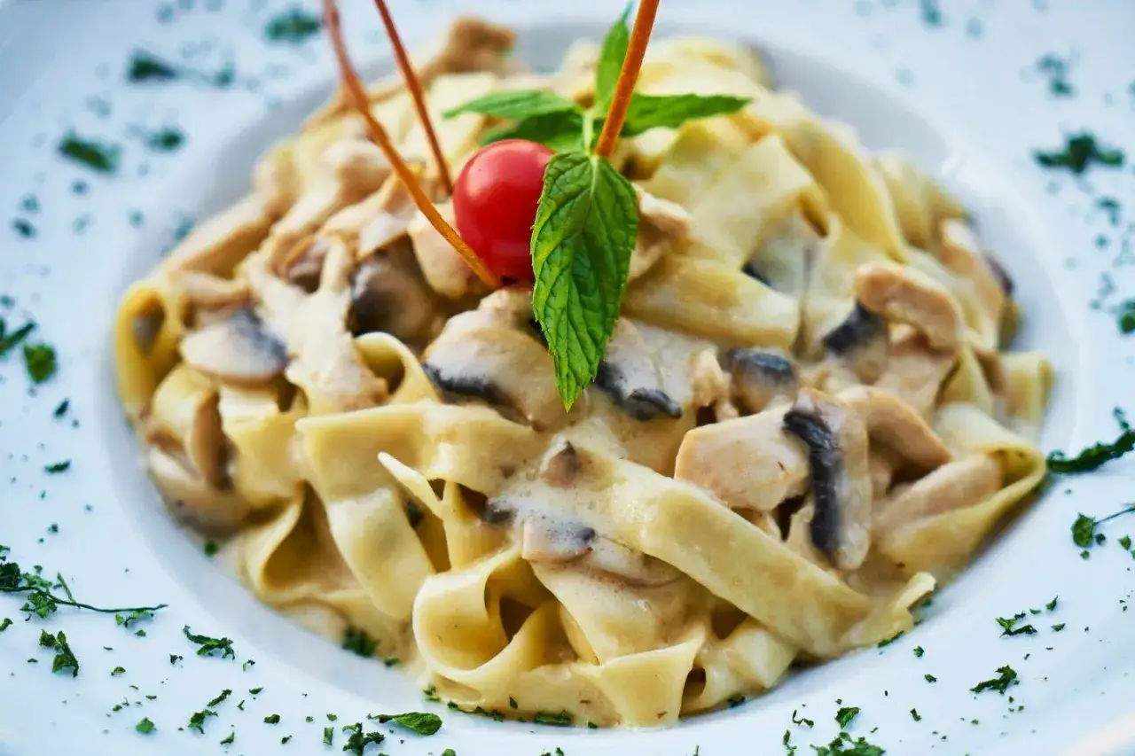 Pyszny kurczak w sosie śmietanowym z grzybami i makaronem tagliatelle, udekorowany pomidorkiem i miętą.